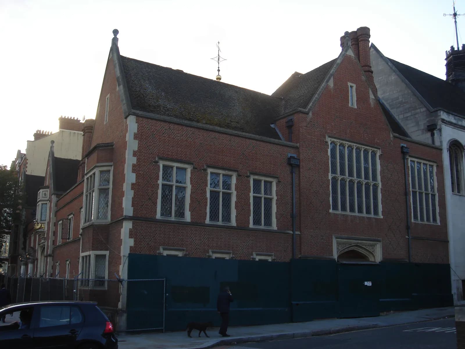 A historic brick building with many windows and a dark roof stands next to a street, with scaffolding or fencing around its base and a car and person on the sidewalk.